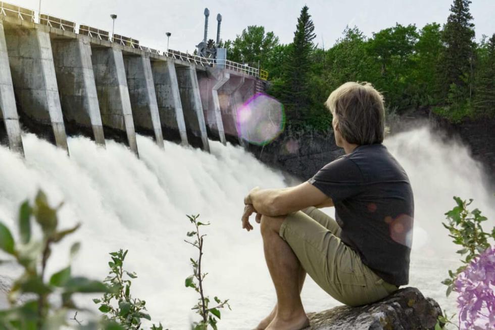 Wasserkraft: Ökostrom aus 100% erneuerbaren Energien Mann sitzt auf einem Stein vor einem Wasserkraftwerk und denkt an die Produktion von erneuerbaren Energien für die Energiewende